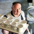 Keith Brymer Jones is pictures holding a tray of white glazed mugs whilst looking up at the camera.